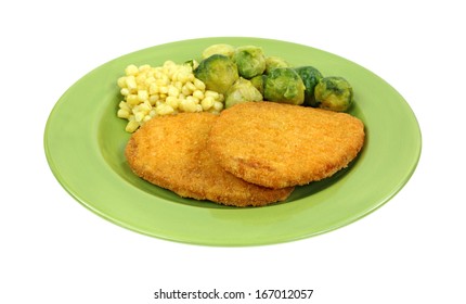 An Angle View Of Two Breaded Veal And Beef Patties With Corn And Brussels Sprouts On A Green Plate Atop White Background.