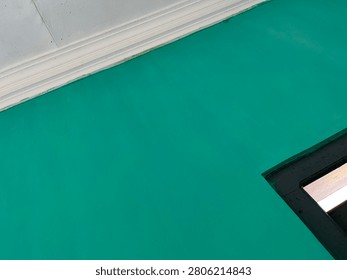 An angled, close-up shot capturing a bold turquoise-green wall background. The striking color is framed by elegant white crown molding at the top and a dark, sharp window structure in the lower right corner, creating a beautiful geometric contrast.