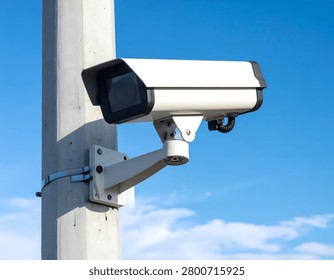 Angled close-up of a white, rectangular security camera mounted on a concrete pole against a blue sky