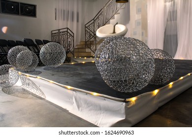 Angled Shot Of A Fashion Show Runway Set Decorated With A White Seat, Led Light Strips And Silver Metal Mesh Balls On A Low Black Stage With Stairs, Folding Chairs And Sheer Drapes In The Background