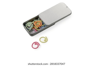 An angled studio product photograph of a rectangular metal tin box with a sliding lid partially closed, accompanied by two loose spiral plastic stitch markers (pink and light green) placed in front of it, filled with an assortment of colorful markers and isolated against a pure white background.