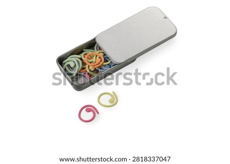 An angled studio product photograph of a rectangular metal tin box with a sliding lid partially closed, accompanied by two loose spiral plastic stitch markers (pink and light green) placed in front of it, filled with an assortment of colorful markers and isolated against a pure white background.