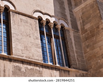 An angled view of elegant arched windows set in a historic stone facade, featuring delicate carved columns and multi-pane leaded glass. Highlights classic Gothic and Renaissance architectural elements, master stonemasonry, and European cultural heritage buildings.