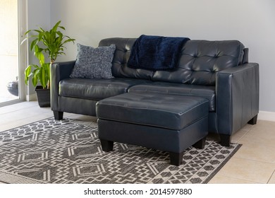 An Angled View Of A Large Blue Moden Styled Love Seat And Ottoman Sofa Made Of Genuine Leather. The Couch Is By A Large Sliding Door And Neutral Colored Tile And Geometric Carpet. 