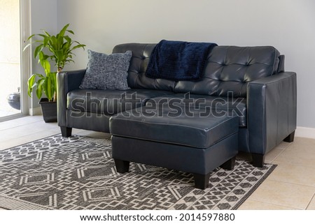 An angled view of a large blue moden styled love seat and ottoman sofa made of genuine leather. The couch is by a large sliding door and neutral colored tile and geometric carpet. 