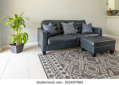 An Angled View Of A  Large Blue Modern Styled Love Seat And Ottoman Sofa Made Of Genuine Leather. The Couch Is By A Large Bright Window And Surrounded With Neutral Colored Tile Floor And Wall. 