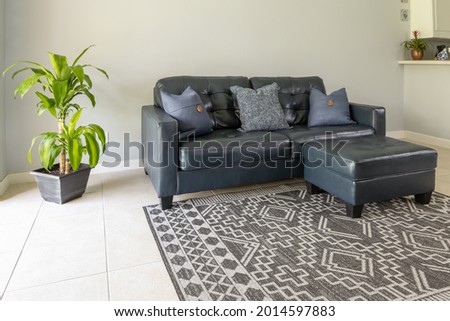 An angled view of a  large blue modern styled love seat and ottoman sofa made of genuine leather. The couch is by a large bright window and surrounded with neutral colored tile floor and wall. 