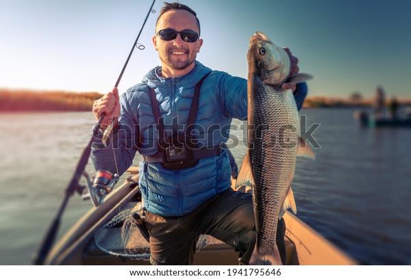 Angler with Asp\
fish. Amateur fisherman holds the asp fish and stands in the boat\
with river on the\
background