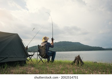 Angler sitting a chair and cast rod fishing at the lake. Fisherman with camping tent and bonfire for cooking on the shore of the lake. Survival concept. - Powered by Shutterstock