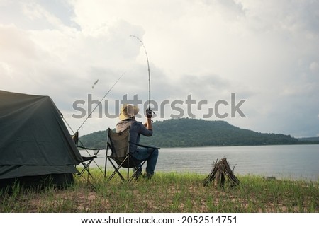 Angler sitting a chair and cast rod fishing at the lake. Fisherman with camping tent and bonfire for cooking on the shore of the lake. Survival concept.