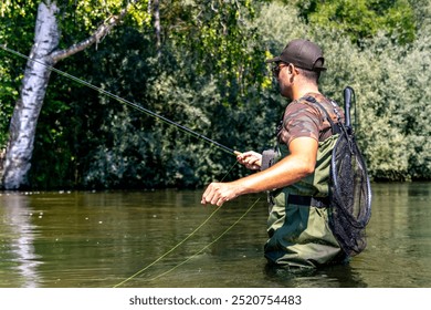 An angler standing in a shallow river, skillfully casting his fly fishing rod, with a net attached to his back and surrounded by lush greenery. - Powered by Shutterstock