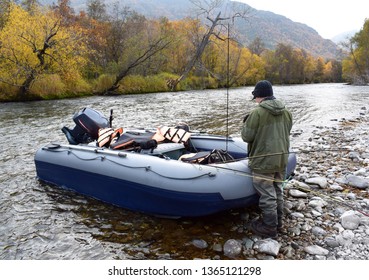 Angler Teen With Fishing Rods Stands On The Rocky Shore Of The Rapid River At The Inflatable Rubber Boat With Motor And Fishing Rods