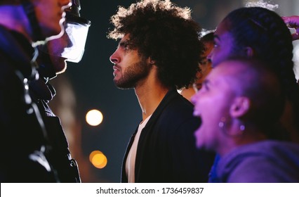 Angry activists protesting against police violence. Young man looking into eyes of a policeman while standing with group of protestors at night. - Shutterstock ID 1736459837