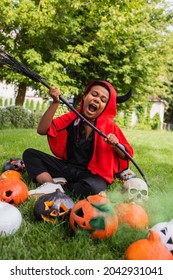 Angry African American Boy In Devil Halloween Costume Screaming While Holding Broom Near Pumpkins And Sitting On Lawn