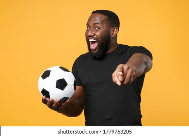 Angry African American Man Football Fan In Black T-shirt Isolated On Yellow Background. Sport Family Leisure Concept. Cheer Up Support Favorite Team With Soccer Ball Pointing Index Finger On Camera