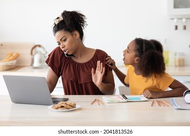 Angry african american teen girl sitting by working mom, asking for attention, kitchen interior. Busy black lady working from home, using laptop and having phone conversation, have no time for kid