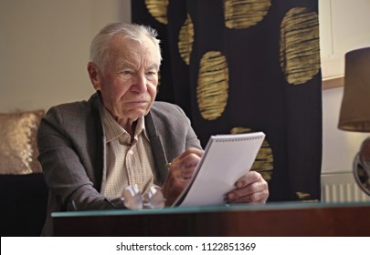 Angry and annoyed grey haired senior man writing notes to the notebook sitting at the table in the living room. Finance, education, leisure activities and retirement planning concept.