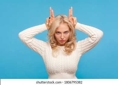 Angry Arrogant Woman With Curly Blond Hair Holding Fingers Above Head Showing Horns, Stubborn Bossy Female Demonstrating Her Temper. Indoor Studio Shot Isolated On Blue Background