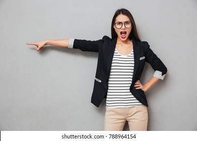 Angry Asian Business Woman In Eyeglasses With Arm On Hip Pointing Away And Looking At The Camera Over Gray Background