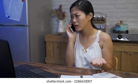 Angry Asian Female Is Pointing At The Monitor And Hitting The Table While Making A Phone Call To Complain About The Incorrect Billing At Home.