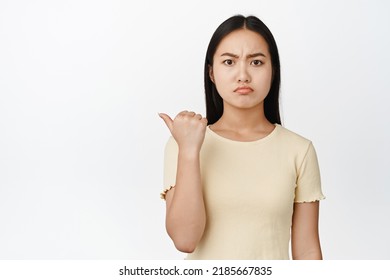 Angry Asian Girl Looks Grumpy Or Jealous, Points Left At Empty Space, White Background