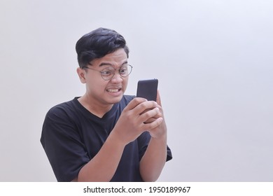 Angry Asian Man In Black T-shirt Wearing Glasses, Getting Mad While Looking At Mobile Phone. Teenager Holding A Cellphone With Arms Raised. Isolated Image On Gray Background