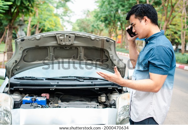 Angry Asian\
man and using mobile phone calling for assistance after a car\
breakdown on street. Concept of vehicle engine problem or accident\
and emergency help from Professional\
mechanic