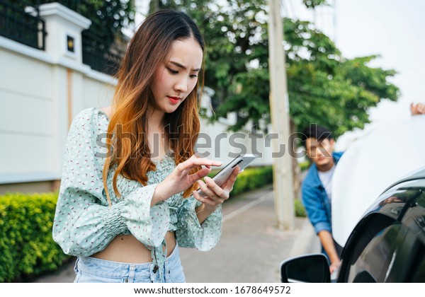 Angry asian woman start calling\
mechanic with phone to come here and fix her car. Young woman using\
mobile phone while looking at broken down car on\
street.
