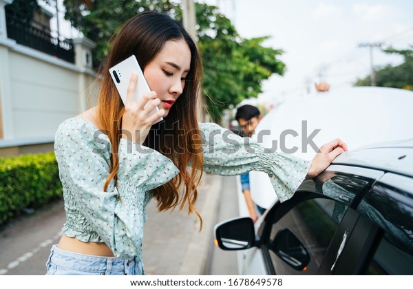 Angry asian woman start calling\
mechanic with phone to come here and fix her car. Young woman using\
mobile phone while looking at broken down car on\
street.