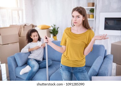 I Am Angry. Beautiful Young Girl Cleaning The Floor And Being Mad At Her Lazy Roommate While She Sitting Texting On The Sofa Instead Of Helping Her