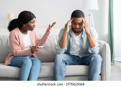 Angry Black Woman With Pregnancy Test Yelling At Stressed Man, Sitting On Couch Together In Living Room. Pregnant African American Lady Fighting With Her Boyfriend. Unwilling Pregnancy Concept