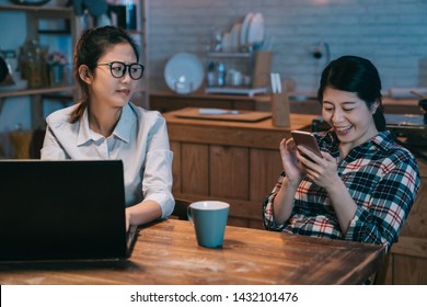 Angry Business Worker Woman Looking At Lazy Mate Playing With The Smart Phone. Annoyed Young Girl Roommate Friend Laughing Loudly Sitting Next To Hard Working Employee In Home Kitchen.