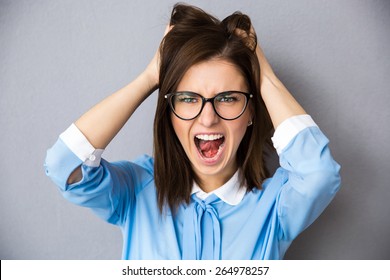 Angry Businesswoman Touching Her Hair And Screaming Over Gray Background. Wearing In Blue Shirt And Glasses. Looking At Camera