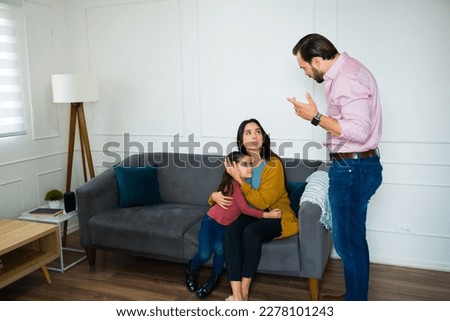 Angry caucasian man screaming to his scared family while the mom is hugging their little daughter 