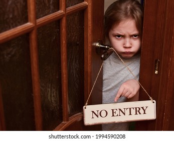 An Angry Child Closes The Door To His Room And Points To A Sign That Says No Entry. The Child Does Not Let Her Parents Into The Room. Upbringing Problems, The Conquest Of Personal Space By Children