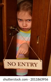 An Angry Child Closes The Door To His Room And Points To A Sign That Says No Entry. The Child Does Not Let Her Parents Into The Room. Upbringing Problems, The Conquest Of Personal Space By Children