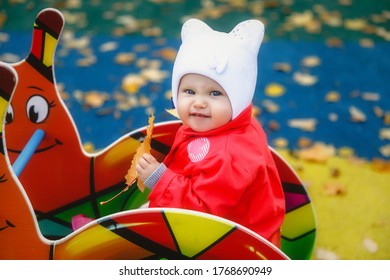 Angry Child In Playground. Pretty Blue-eyed One-year-old Girl In Red Coat And White Hat. Autumn Yellow Leaf In Hands. Charming Child Shows Emotions In Close-up. Concept Of Healthy Child And Parenting