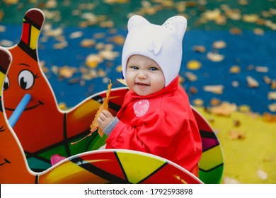 Angry Child In Playground. Pretty Blue-eyed One-year-old Girl In Red Coat And White Hat. Autumn Yellow Leaf In Hands. Charming Child Shows Emotions In Close-up. Concept Of Healthy Child And Parenting