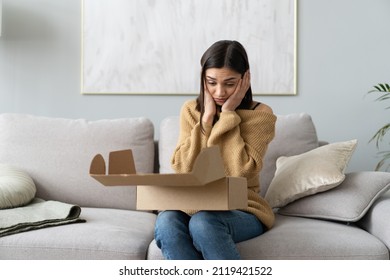 Angry confused woman unpacking parcel, wrong or broken online store order, sitting on couch at home, dissatisfied female looking in cardboard box, bad delivery service, displeased by post shipping