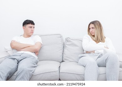 Angry Couple Sitting Separated In The Sofa Looking At Each Other. Mad Couple After A Relationship Conflict. Young Couple At Home. Heterosexual 18-20 Years Old Couple.