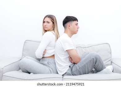 Angry Couple Sitting In The Sofa Back To Back With Crossed Arms. Woman Looking At Camera. Young Couple At Home. Heterosexual 18-20 Years Old Couple.