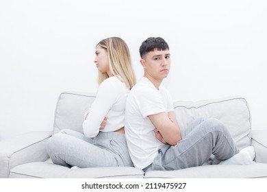 Angry Couple Sitting In The Sofa Back To Back With Crossed Arms. Man Looking At Camera. Mad Couple After A Relationship Conflict.  Young Couple At Home. Heterosexual 18-20 Years Old Couple.