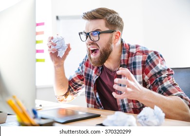 Angry crazy modern designer in glasses with beard yelling and crumpling paper on his workplace