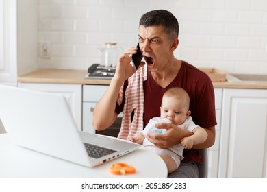 Angry Dark Haired Handsome Man Wearing T Shirt With Towel On His Shoulder, Talking Via Cell Phone And Screaming With Aggressive Expression, Sitting At Table With Laptop, Holding Baby Girl In Hands.