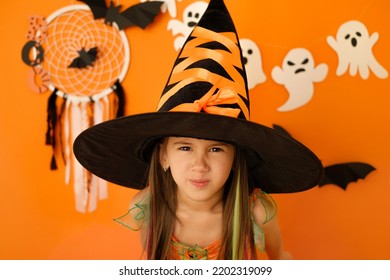 An angry dissatisfied girl in the guise of a witch looks into the camera making a face. Halloween decor with ghosts on an orange studio background - Shutterstock ID 2202319099
