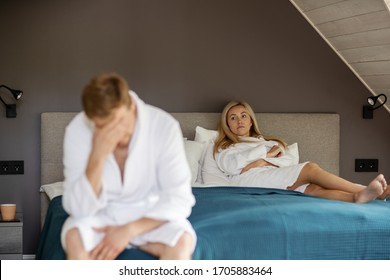 Angry dissatisfied woman in white bathrobe lying on bed while her defocused depressed husband or lover rubbing forehead in foreground