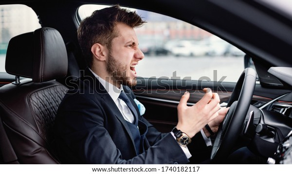 Angry Driver Concept. Aggressive\
mad man in suit driving car in urban city, angrily\
gesturing
