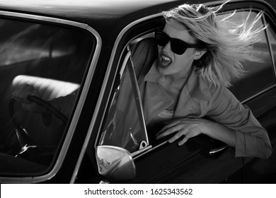 Angry driving blonde woman with black sunglasses. Windy hair. Black and white photo.