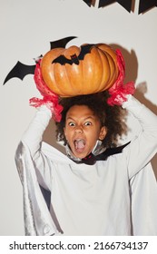 Angry Emotional African Boy In Cape And Red Gloves Holding Large Pumpkin Above Head And Screaming At Camera