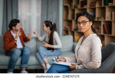 Angry Emotional Young European Couple Quarreling At Meeting With African American Psychologist In Office Clinic Interior. Family Psychological Therapy, Mental Treatment, Professional Help And Business